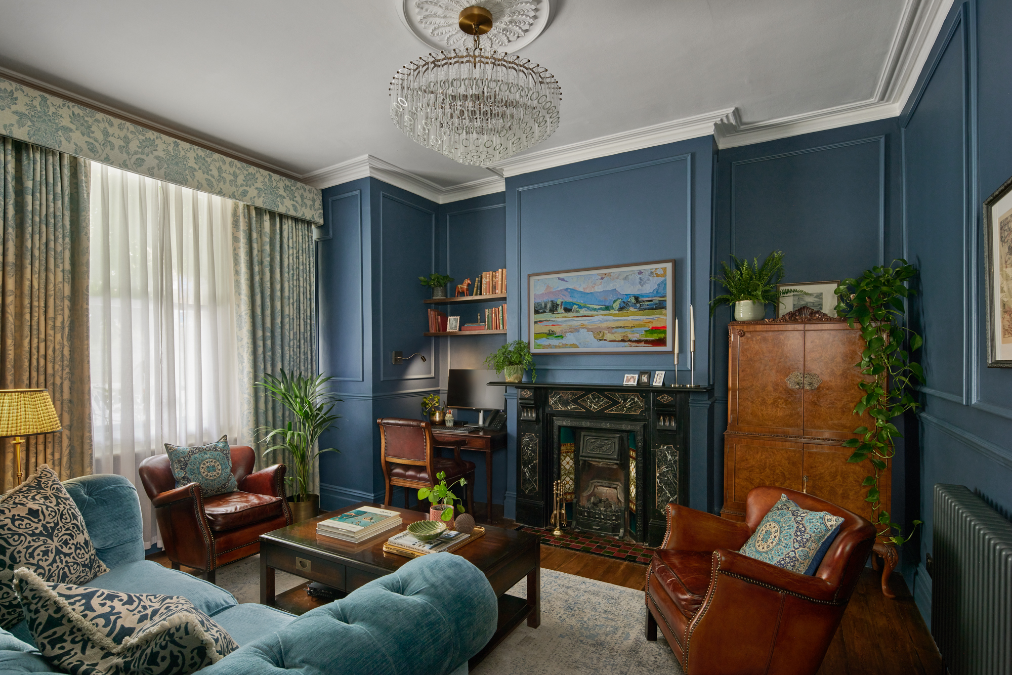 Home 5 A elegant living room in Hammersmith & Fulham designed by Maureen Gomez Interiors, with blue panelled walls, a chandelier, and a Victorian fireplace. The room features a blue sofa, brown leather chairs, a small desk with a computer, bookshelves, and plants. A large painting and patterned curtains add to the decor.
