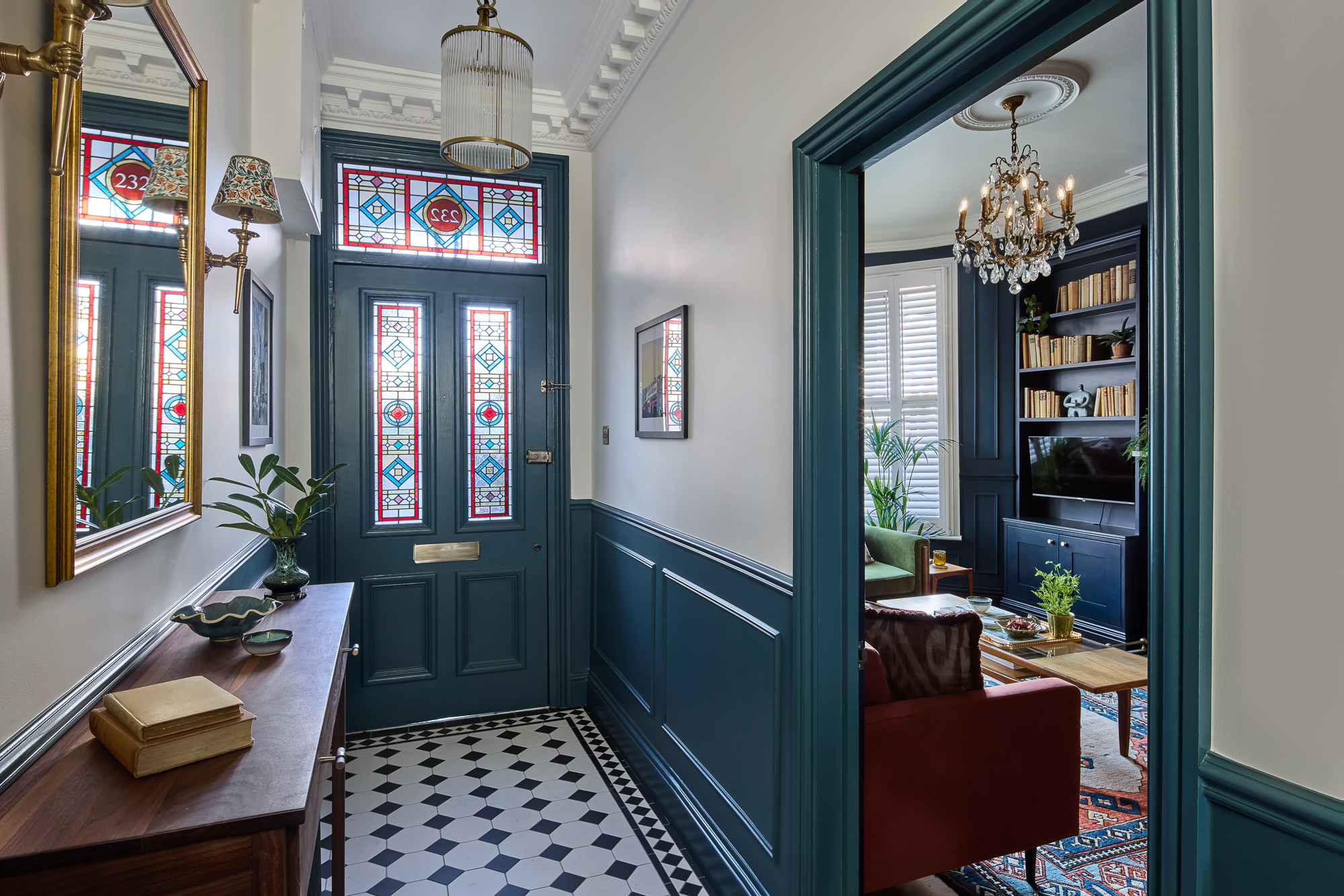 Home 8 A bright hallway with blue wainscoting, a chequered tiled floor, and a stained-glass front door. The hallway leads to a cosy living room with a chandelier, bookshelves, plants, and a red sofa.