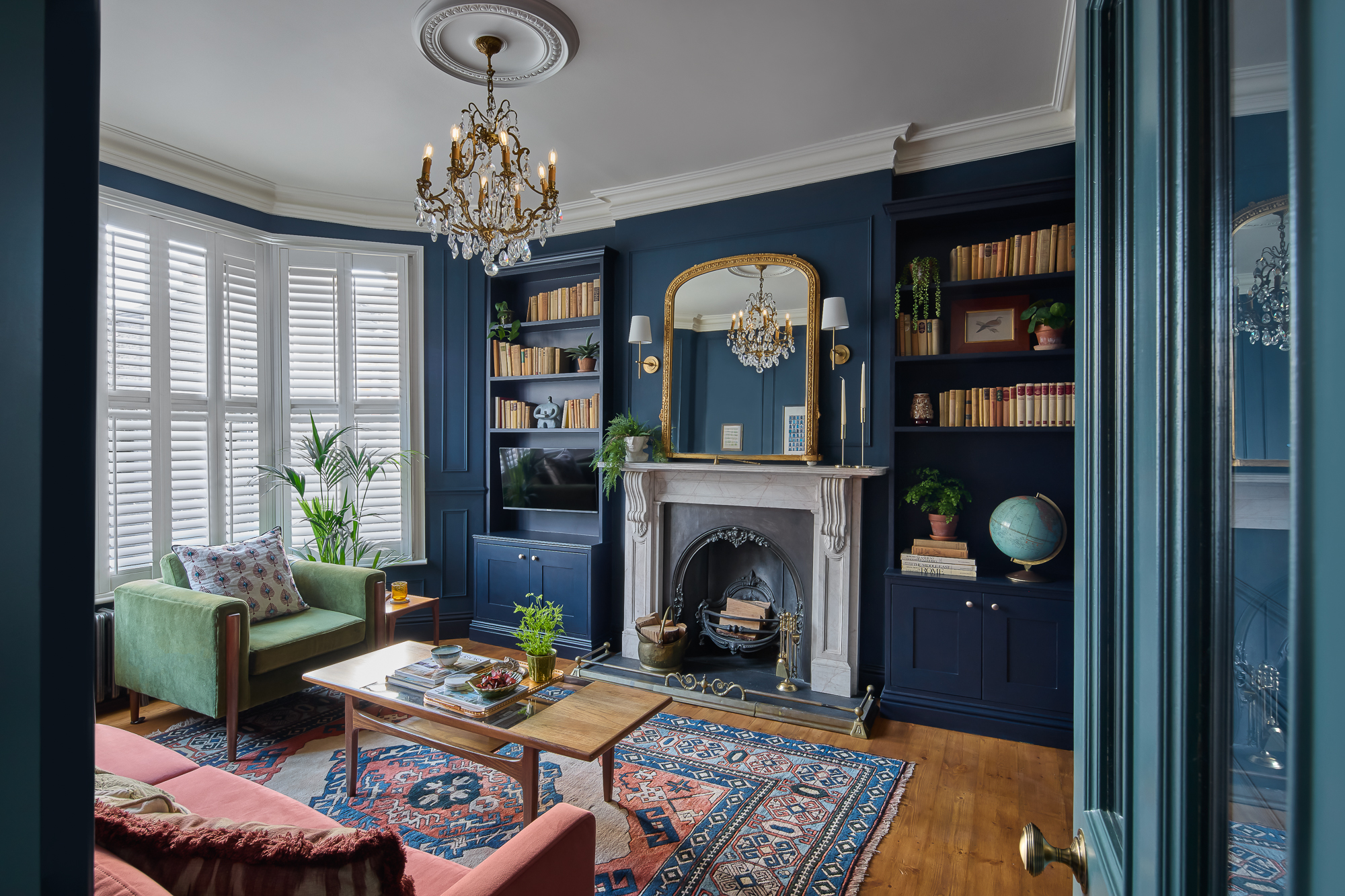 Home 1 A stylish living room with navy blue walls, a white fireplace, bookshelves, a green armchair, a pink sofa, a coffee table, houseplants, a globe, and a chandelier. Large windows have white shutters and light fills the room.