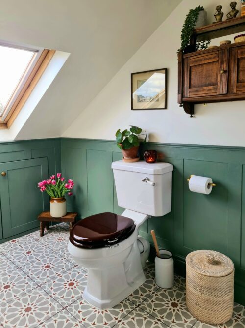 A cosy bathroom designed by Maureen Gomez Interiors, features a white toilet with a dark brown seat, set against green panel walls. A roof window lets in natural light. There's a wicker basket, a small wooden stool with a vase of pink tulips, and a vintage wall cabinet with plants and decor.
