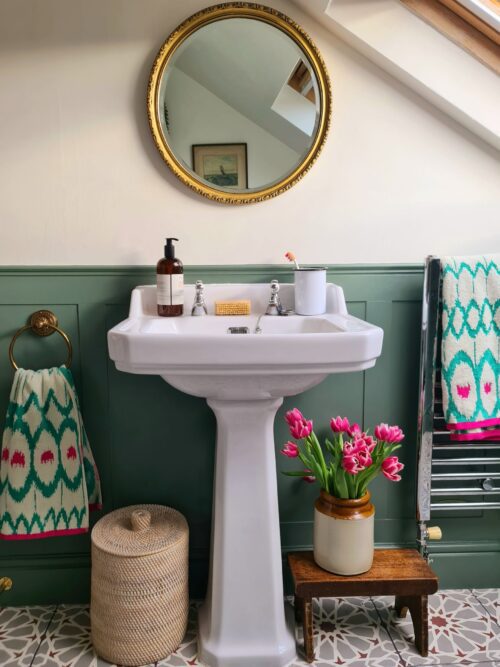 A bathroom designed by Maureen Gomez, Interiors features a white pedestal sink with a round mirror above. A bottle of soap and a cup with a toothbrush sit on the sink. Pink tulips are in a vase on a small stool. There's a towel ring and a towel rail with a colourful towel nearby.