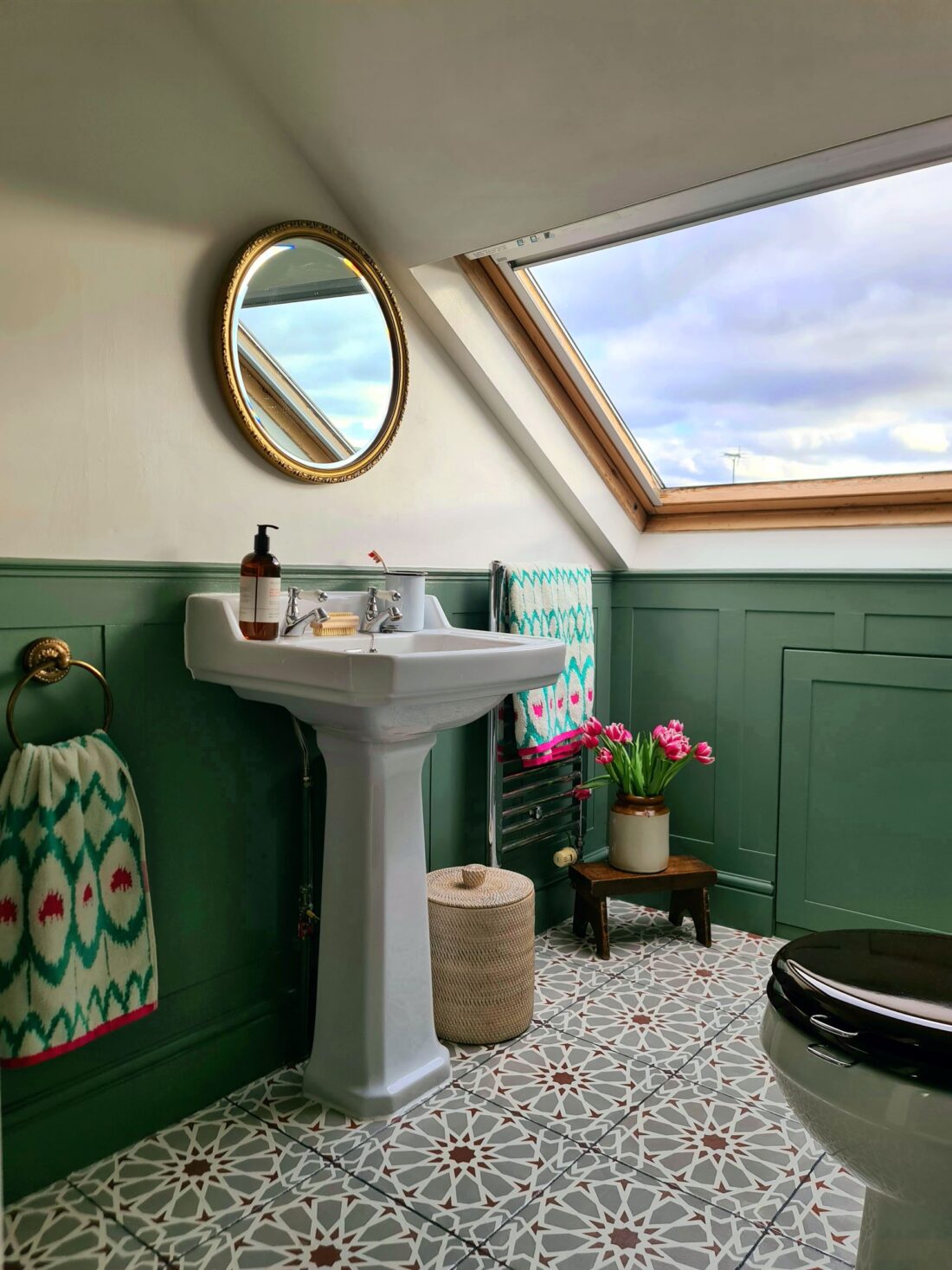 A bathroom designed by Maureen Gomez Interiors, with a white pedestal sink, round mirror, and skylight. It features patterned floor tiles, green wall panelling, a wicker basket, a plant, and colourful towels. A soap dispenser sits on the sink, and pink tulips add a decorative touch.