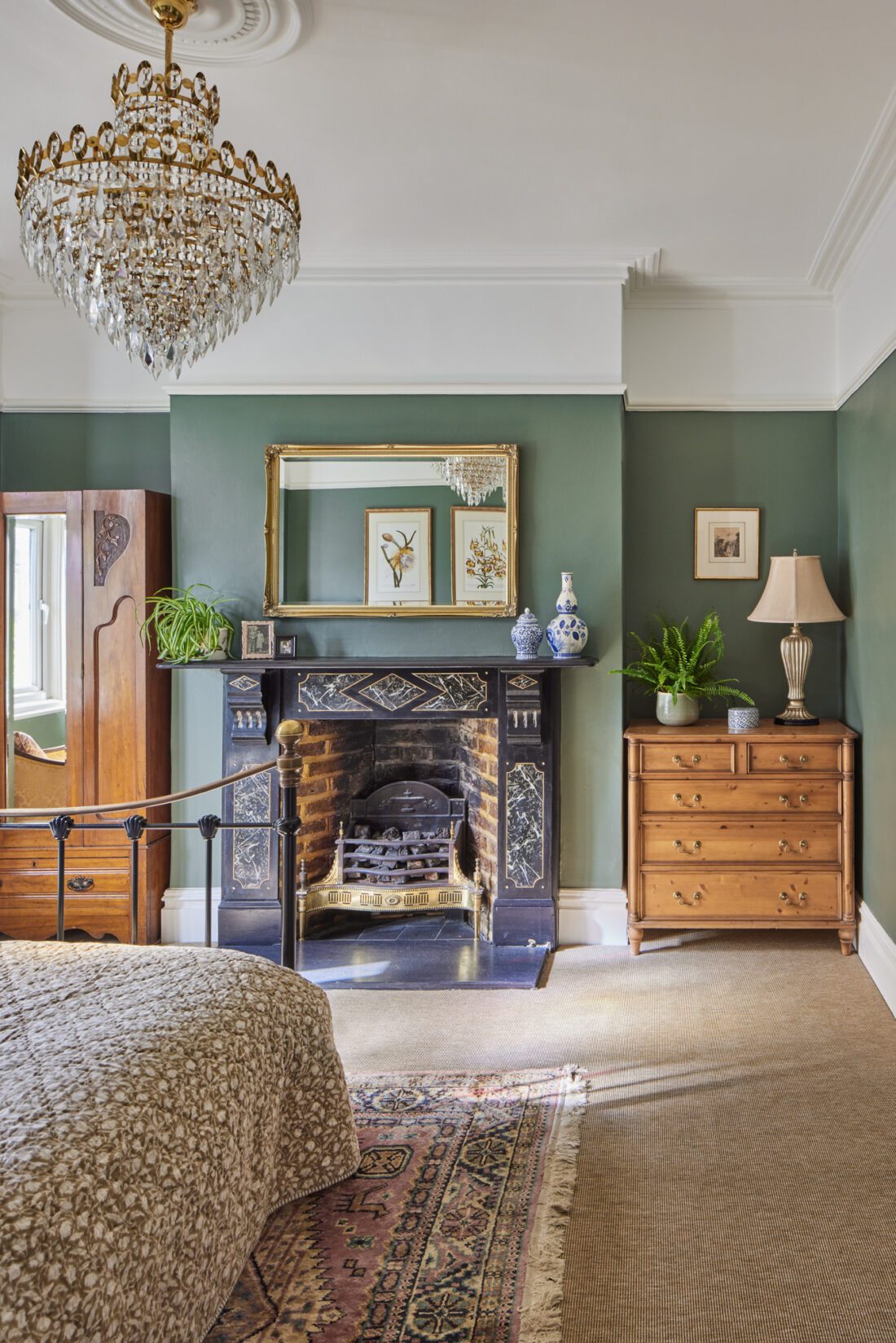 An elegant bedroom with green walls designed by Maureen Gomez Interiors, featuring a chandelier and a decorative fireplace. There's a bed with patterned bedding, an antique wooden wardrobe, a chest of drawers with a lamp and plant, and a mirror above the fireplace reflecting wall art. Natural light fills the room.