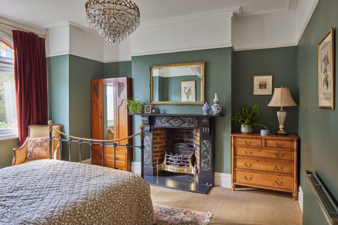 An elegant bedroom with green walls designed by Maureen Gomez Interiors, featuring a metal-framed bed, an ornate black fireplace with a mirror above, an antique wooden wardrobe, and a chest of drawers. A chandelier hangs from the ceiling, and a large window is dressed with red curtains.