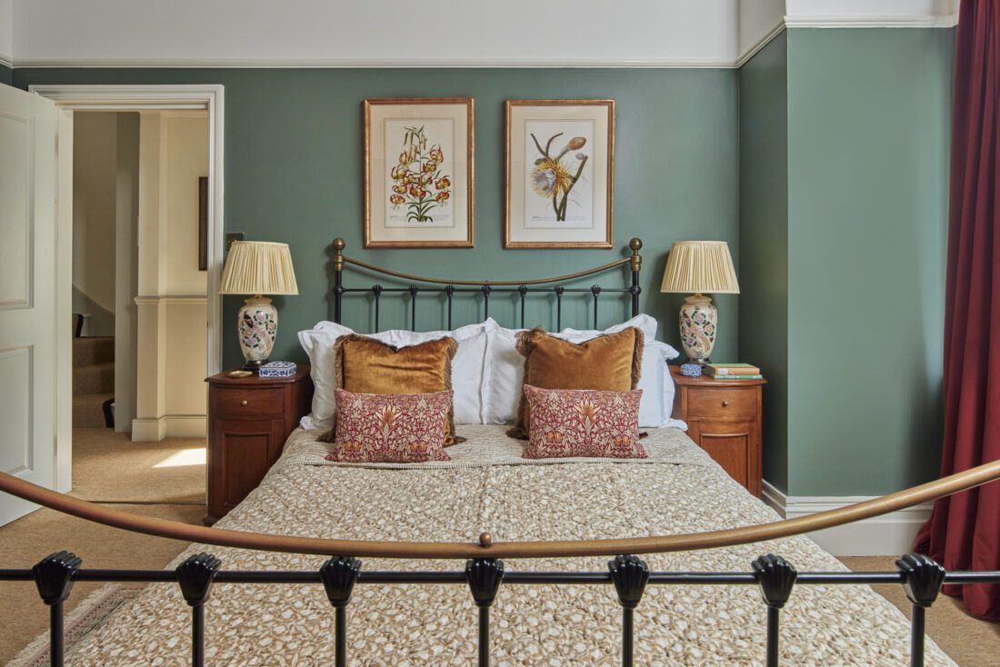 An elegant bedroom designed by Maureen Gomez Interiors, with a metal bed frame, adorned with a patterned bedspread and decorative pillows. Two bedside tables with lamps flank the bed, and floral artwork hangs on green walls. A partially open door leads to a hallway.