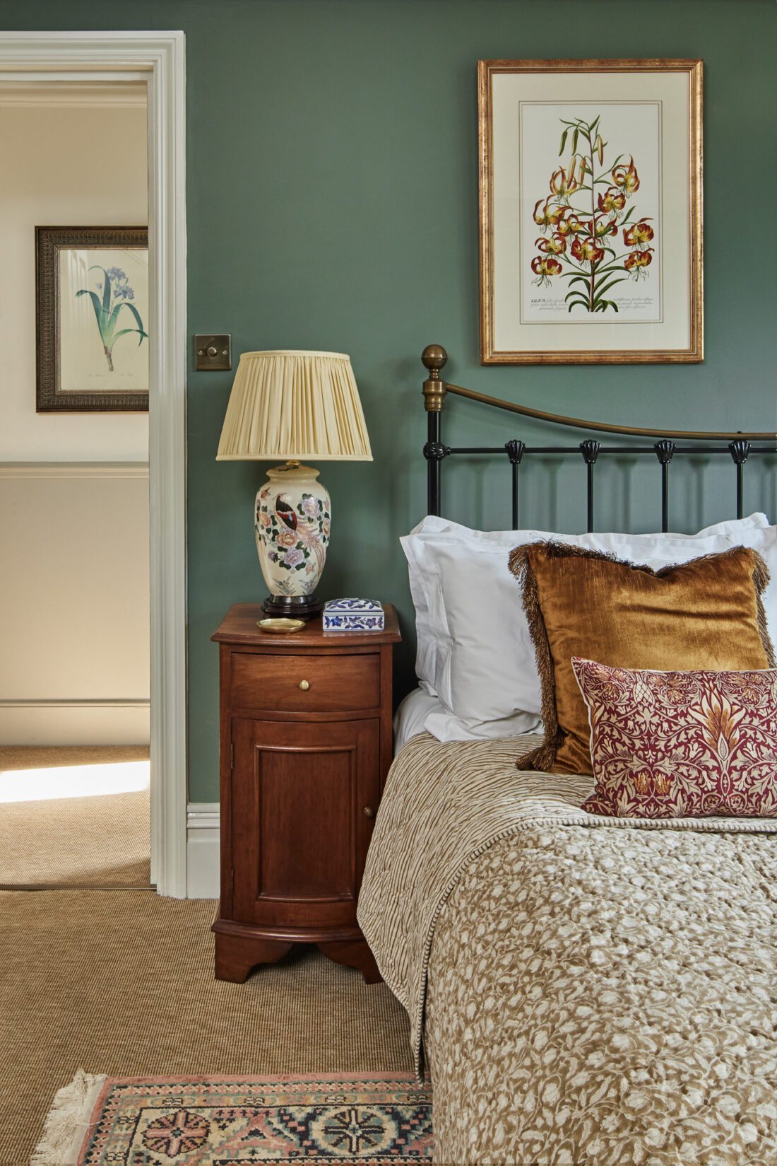 An elegant bedroom with a green wall designed by Maureen Gomez Interiors, features a black metal bed with white sheets and mustard pillows. A wooden nightstand holds a decorative lamp and book. Floral artwork hangs above, and a patterned rug lies on the carpeted floor. An open door leads to another room.