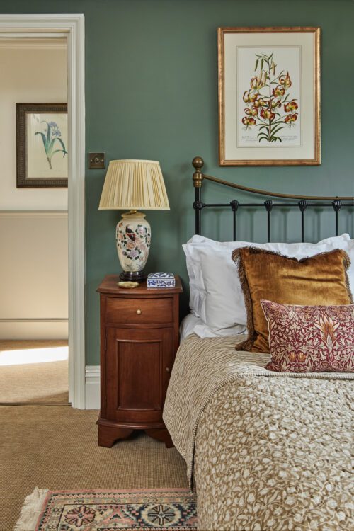 An elegant bedroom with a green wall designed by Maureen Gomez Interiors, features a black metal bed with white sheets and mustard pillows. A wooden nightstand holds a decorative lamp and book. Floral artwork hangs above, and a patterned rug lies on the carpeted floor. An open door leads to another room.