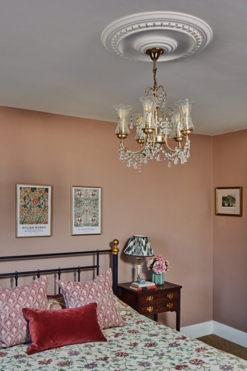 A cosy bedroom designed by Maureen Gomez Interiors, with a floral-patterned bedspread and a black metal bed frame. A side table with a pleated striped lampshade and flowers stands nearby. Two vintage-style framed prints hang on the pink-beige walls, and a crystal chandelier is centred on the ceiling.