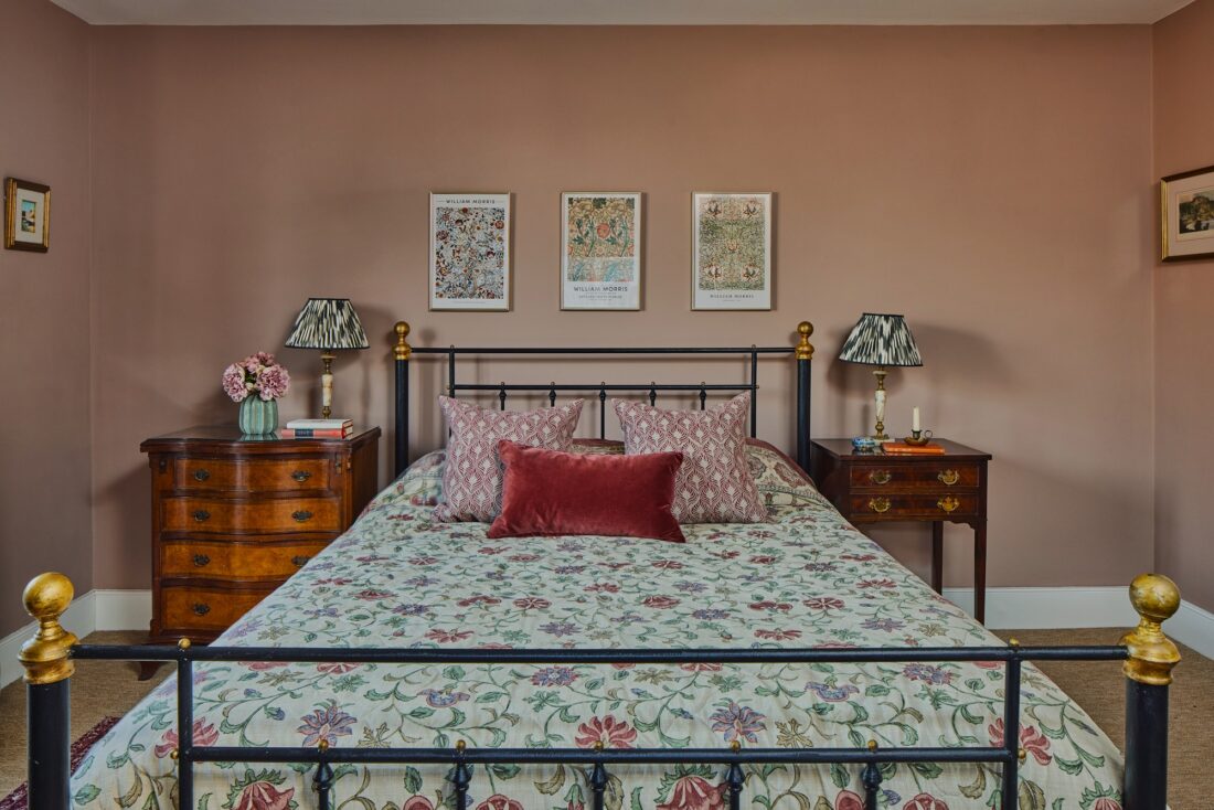 A cosy bedroom designed by Maureen Gomez Interiros, with a floral-patterned bedspread and decorative pillows on a black metal bed. The walls are painted pink-beige with framed artwork above the bed. Wooden nightstands with lamps and a vase of flowers are on each side.