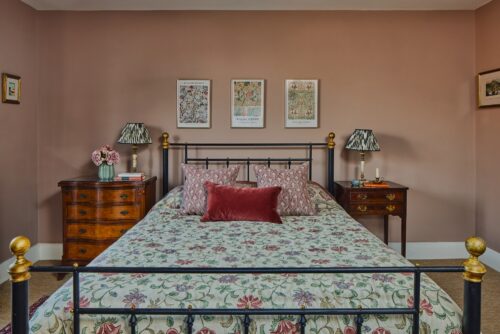 A cosy bedroom designed by Maureen Gomez Interiros, with a floral-patterned bedspread and decorative pillows on a black metal bed. The walls are painted pink-beige with framed artwork above the bed. Wooden nightstands with lamps and a vase of flowers are on each side.