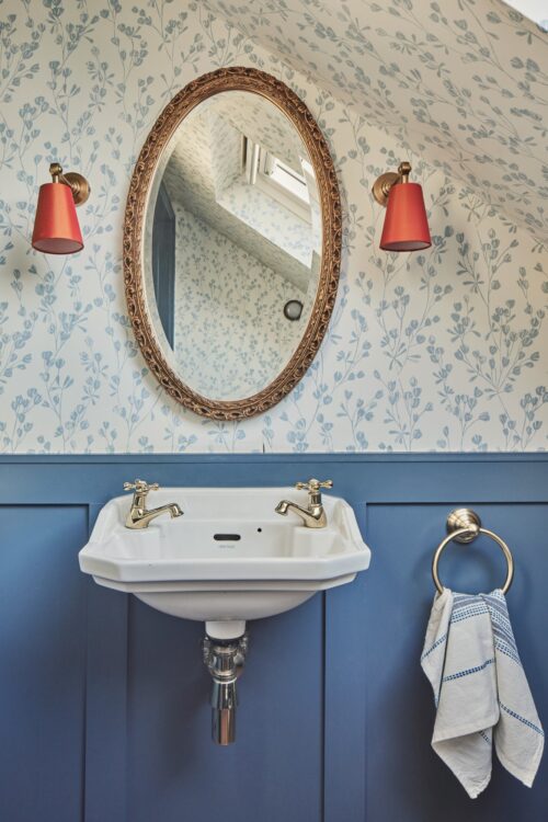 A small bathroom designed byb Maureen Gomez Interiors, features a heritage style basin with gold taps, set against blue panelling. Above, an oval mirror hangs between two orange wall lights. The walls are adorned with floral wallpaper, and a white towel hangs on a gold towel ring.