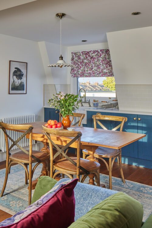 A cosy kitchen and dining area designed by Maureen Gomes Interiors, with a wooden table, four chairs, and a green sofa with colourful cushions. On the tables sits a vase of flowers and a bowl of apples. The kitchen has blue cabinets and a floral Roman shade on the window. A framed picture is on the wall.