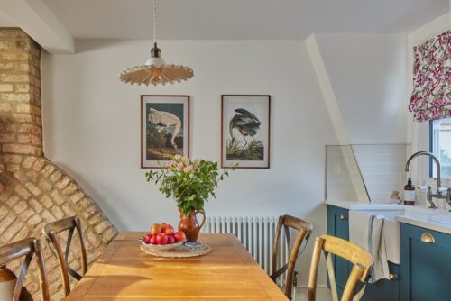 A cosy kitchen designed by Maureen Gomez Interiors, with a wooden table, chairs, and a brick accent wall. The table has a vase with flowers and a bowl of apples. Above, a pendant light hangs, and two framed bird prints adorn the white wall. Blue cabinets are by the sink.