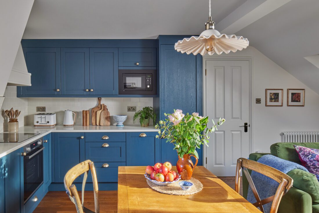 A cosy kitchen-dining room designed by Maureen Gomez Interiors, with blue cabinets and a wooden table and chairs. Fresh flowers and apples sit on the table. A microwave is built into the cabinets, and a frilled pendant light hangs above the table. Plants and framed art add warmth to the space.