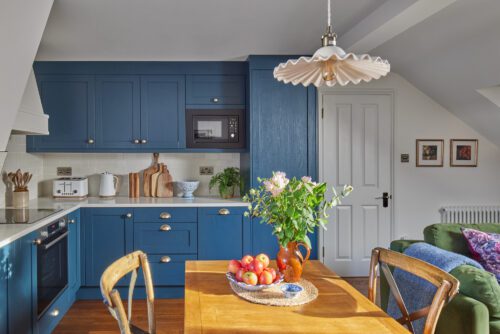 A cosy kitchen-dining room designed by Maureen Gomez Interiors, with blue cabinets and a wooden table and chairs. Fresh flowers and apples sit on the table. A microwave is built into the cabinets, and a frilled pendant light hangs above the table. Plants and framed art add warmth to the space.