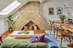 E-design Interior Design UK 3 A cosy loft, living area with a slanted roof and brick arch fireplace, designed by Maureen Gomez Interiors. It features a green sofa, a brass and marble coffee table with decor, a dining table with rustic wooden chairs, artwork on the walls, and a skylight illuminating the space.