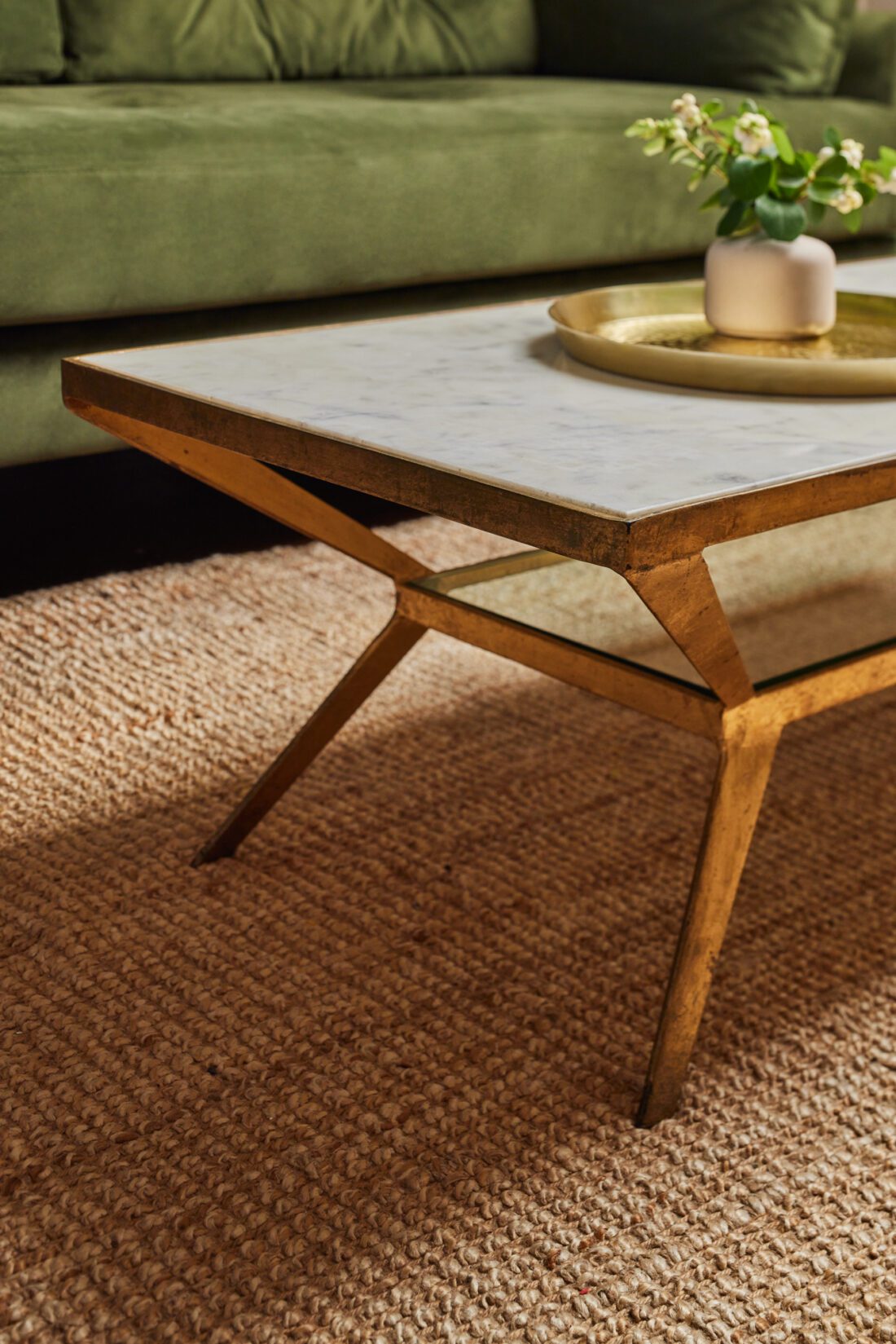A modern coffee table with a marble top and gold frame is situated on a textured rug in a living room design by Maureen Gomez Interiors. On the table, there's a gold tray with a small white vase containing greenery. A green sofa is visible in the background.