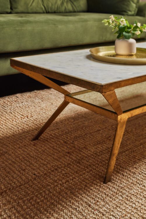 A modern coffee table with a marble top and gold frame is situated on a textured rug in a living room design by Maureen Gomez Interiors. On the table, there's a gold tray with a small white vase containing greenery. A green sofa is visible in the background.