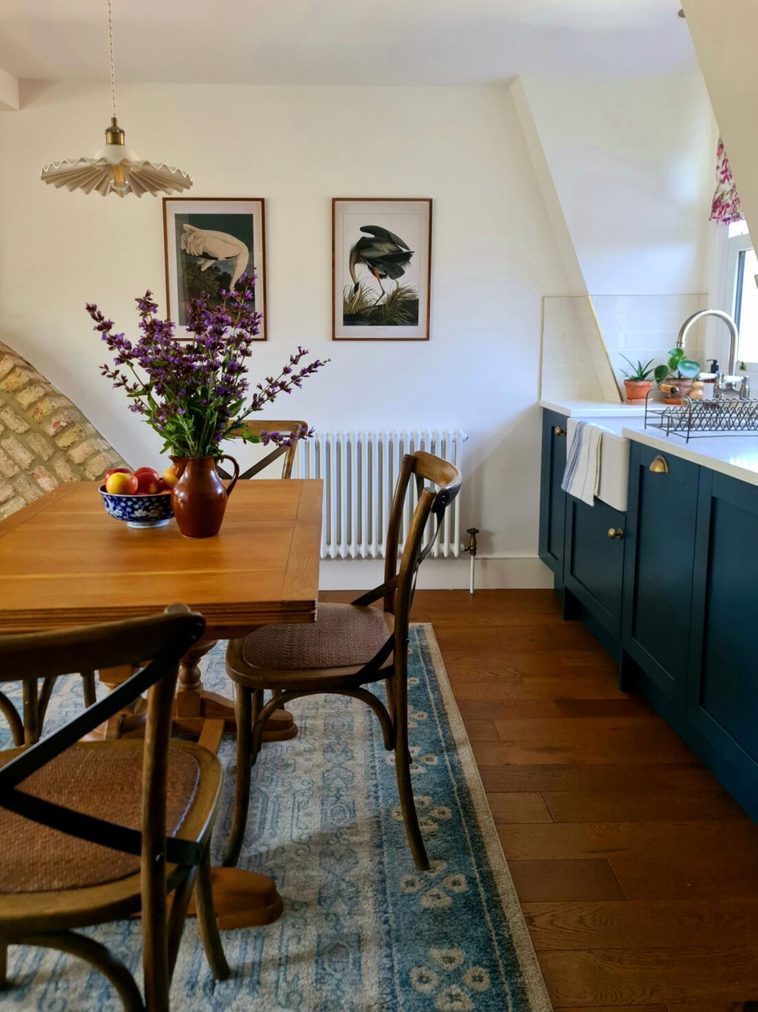 A cosy kitchen-dining area designed by Maureen Gomez Interiors, with a wooden table set with chairs and a vase of purple flowers. A frilly pendant hangs over the table. The kitchen features dark blue cabinets, a white countertop, and a window. Two framed bird prints are on the wall, and a blue patterned rug is on the floor.