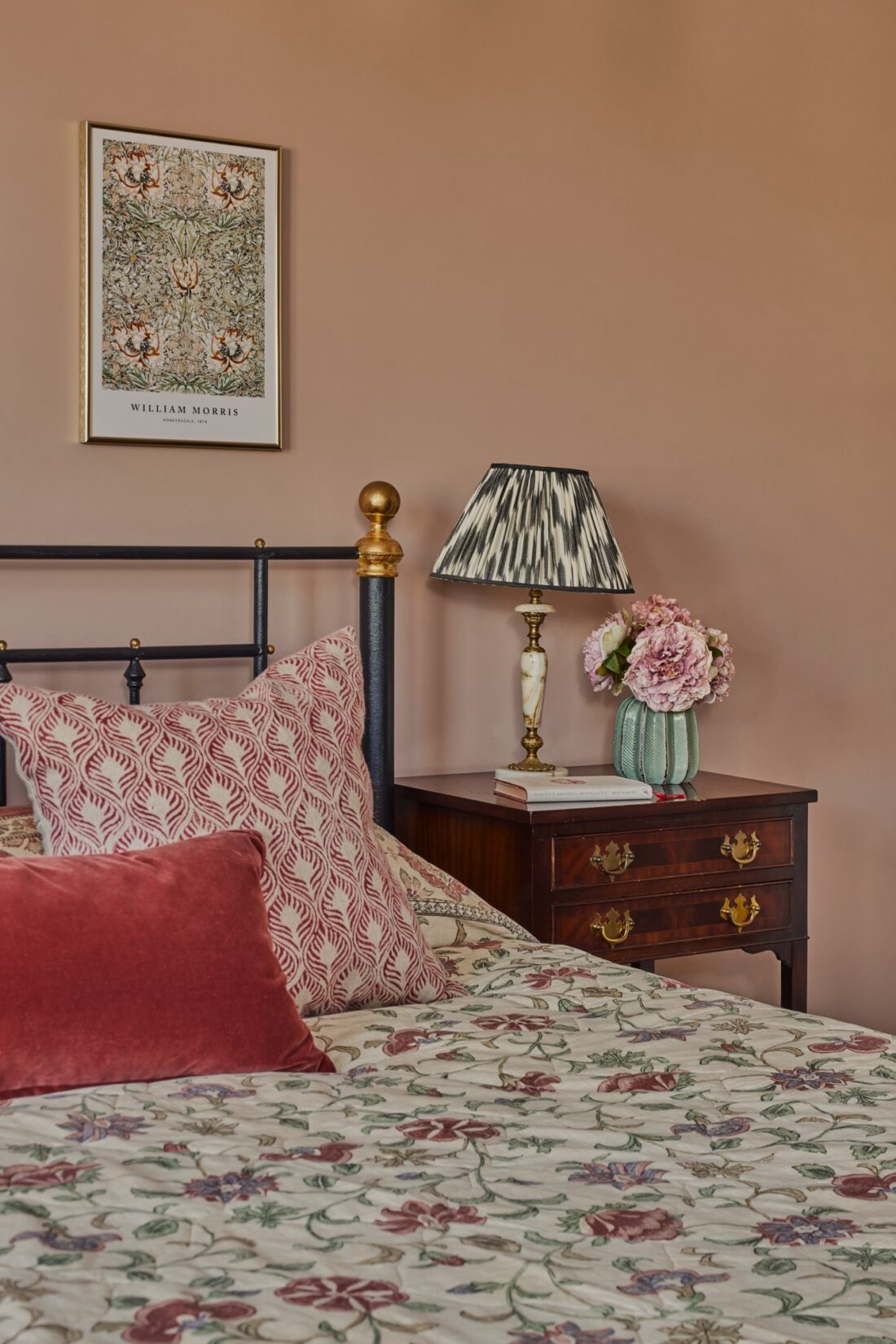 A cosy bedroom designed by Maureen Gomez Interiors, features a bed with a floral bedspread and pink pillows. An antique wood side table holds a patterned lamp and a vase with pink flowers. A framed William Morris print hangs on the pink-beige wall.