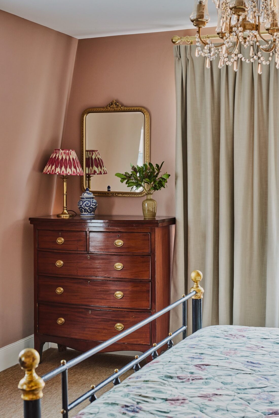 A cosy bedroom designed by Maureen Gomez Interiors, with an antique wood dresser holding a lamp and a vase with greenery. A gold-framed mirror hangs above. The room has pink-beige walls, beige linen curtains, a chandelier, and a bed with a floral-patterned bedspread.