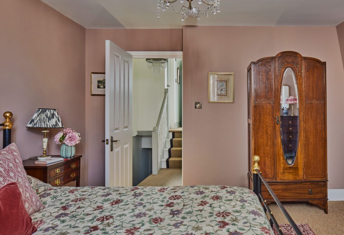 A cosy bedroom designed by Maureen Gomez Interiors, with an antique wood dresser holding a lamp and a vase with greenery. A gold-framed mirror hangs above. The room has pink-beige walls, beige linen curtains, a chandelier, and a bed with a floral-patterned bedspread.