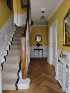 A hallway designed by Maureen Gomez Interiors, with yellow walls and white panelling on the lower half of the walls. It features oak herringbone flooring and a staircase with a beige stair runner and dark trim. A round mirror and small table with flowers are at the end. A lantern-style light pendant hangs from the ceiling, and a radiator cover is on the right wall.