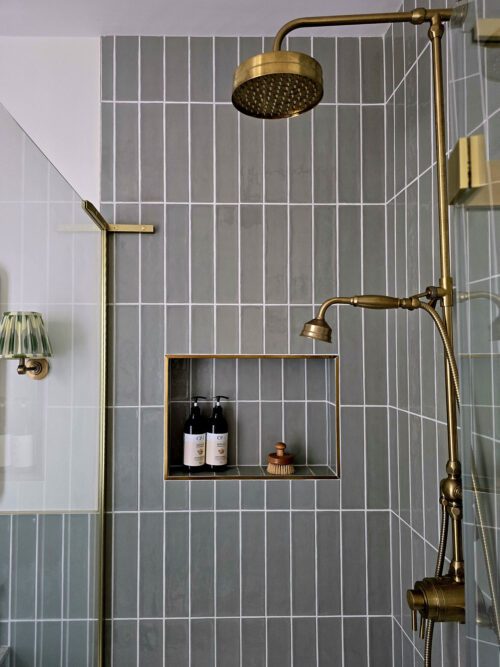A modern bathroom designed by Maureen Gomez Interiors with blue-green vertical tiles. It features a brass shower head and adjustable handheld shower. A niche holds two shampoo bottles and a brush. A glass shower door and a vintage-style wall light are also visible.