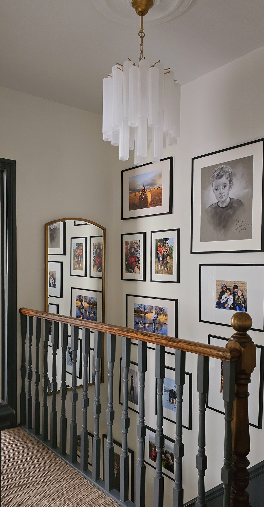 A landing designed by Maureen Gomez Interiors, with a wooden railing displays various framed family photos and portraits on the walls. A tall rectangular mirror reflects the gallery, and a white tiered Art Deco style chandelier hangs from the ceiling above.