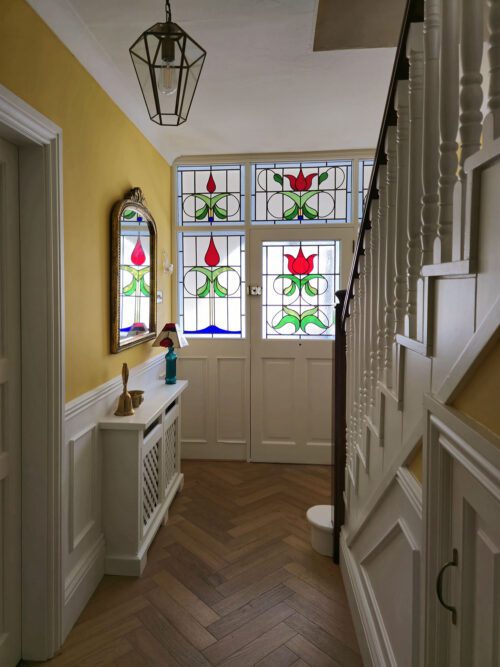 A hallway designed by Maureen Gomez Interiors, with yellow walls, white panelling and wooden herringbone flooring. It features a stained glass door with traditional Edwardian designs. A mirror and small shelf are on the left side, and a staircase with white railing is on the right. A lantern style pendant light hangs from the ceiling.