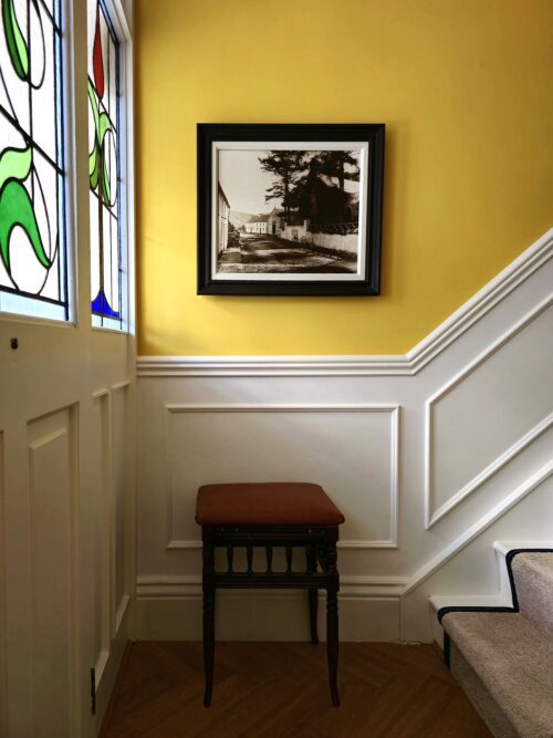 A cosy corner in a hallway designed by Maureen Gomez Interiors, with a yellow wall and white panelling. A framed black-and-white photograph hangs above a small wooden stool. Natural light filters through a stained glass window on the left.
