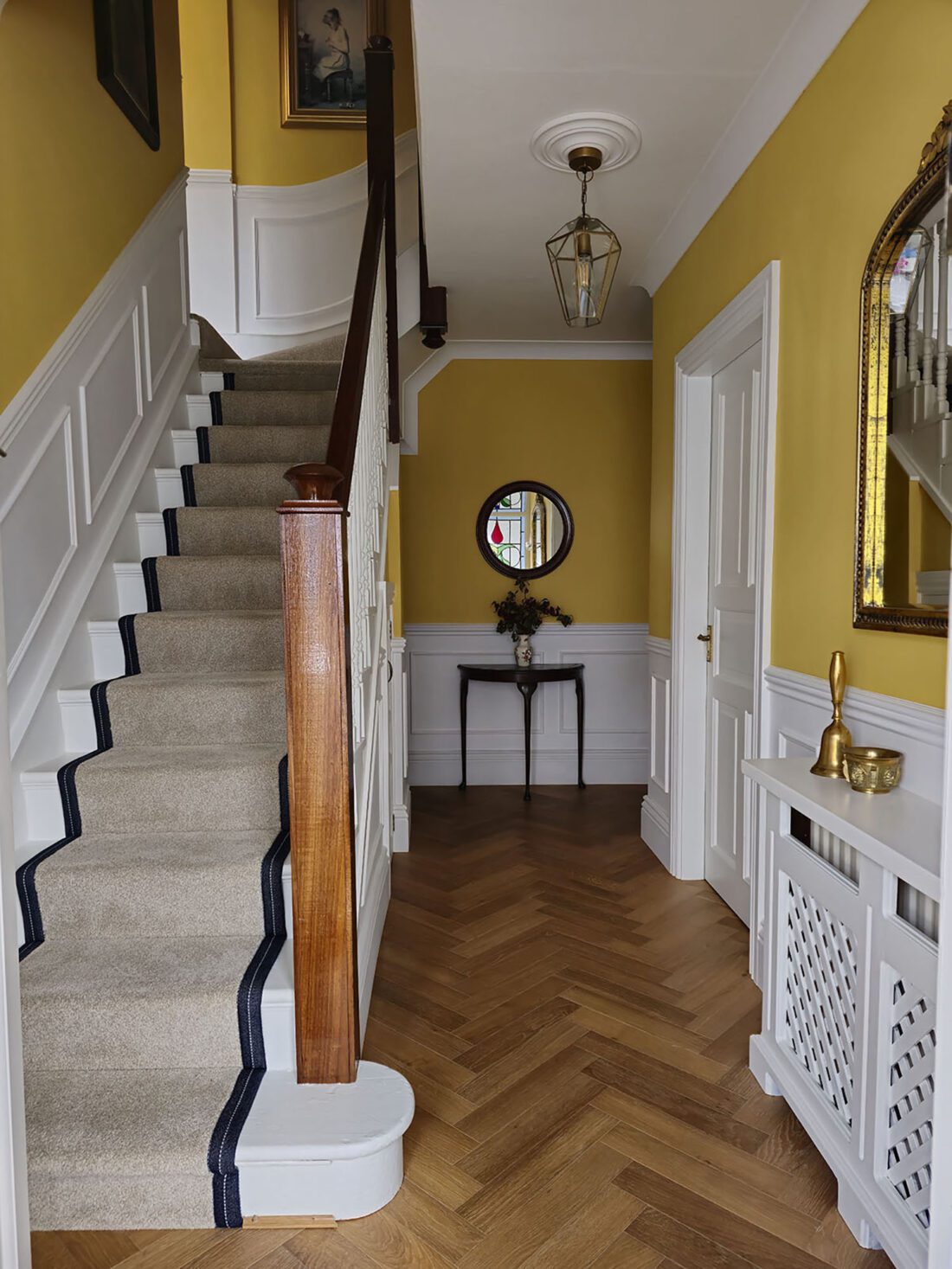 A hallway designed by Maureen Gomez Interiors, with yellow walls and white panelling on the lower half of the walls. It features a staircase with a beige stair runner and dark trim. A round mirror and small table with flowers are at the end. A lantern-style light pendant hangs from the ceiling, and a radiator cover is on the right wall.