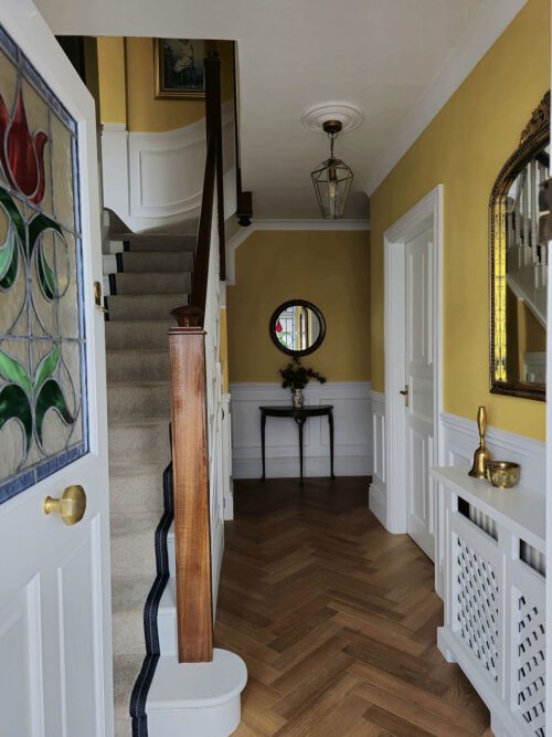 A hallway designed by Maureen Gomez Interiors, with yellow walls and white panelling features a wooden staircase, herringbone wood flooring, and a stained glass door. A round mirror and a small table with a vase of flowers are visible. A lantern style pendant light hangs from the ceiling.