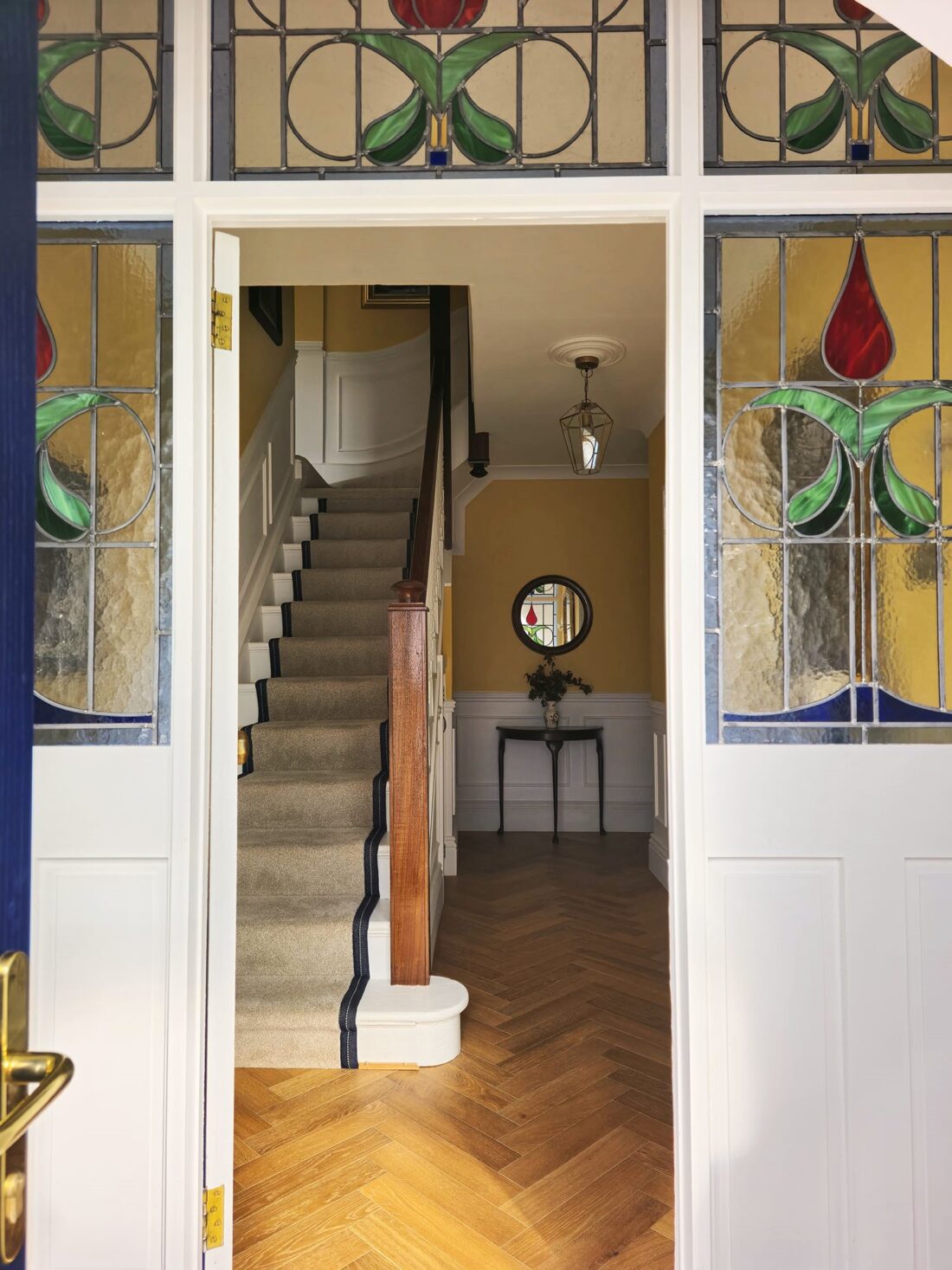 Open door revealing an entryway with Edwardian stained glass panels, featuring a staircase with a dark wooden railing, designed by Maureen Gomez Interiors. The hall has herringbone wood floors, a small table with a vase of flowers, and a mirror on the wall. The upper walls are painted yellow with white panelling below.