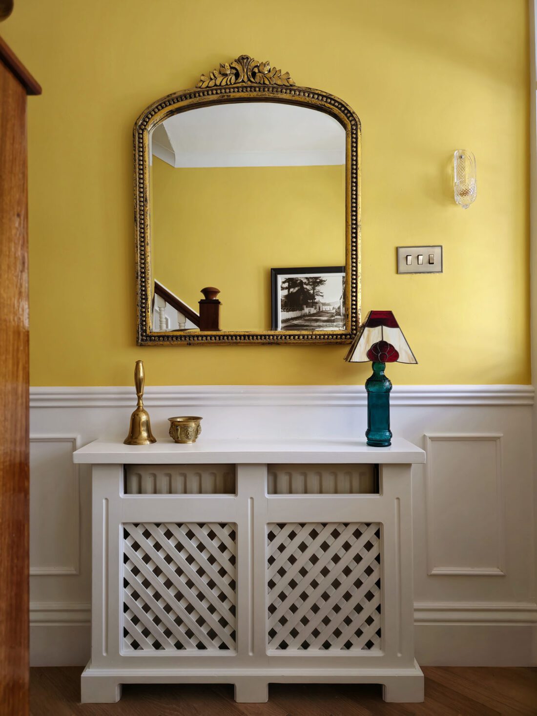 A hallway designed by Maureen Gomez Interiors, with a yellow wall and white panelling, featuring a decorative mirror with a gold frame above a white lattice radiator cover. On top sits a blue lamp with a red lampshade and a small brass candle holder. A framed black and white picture is in the background.