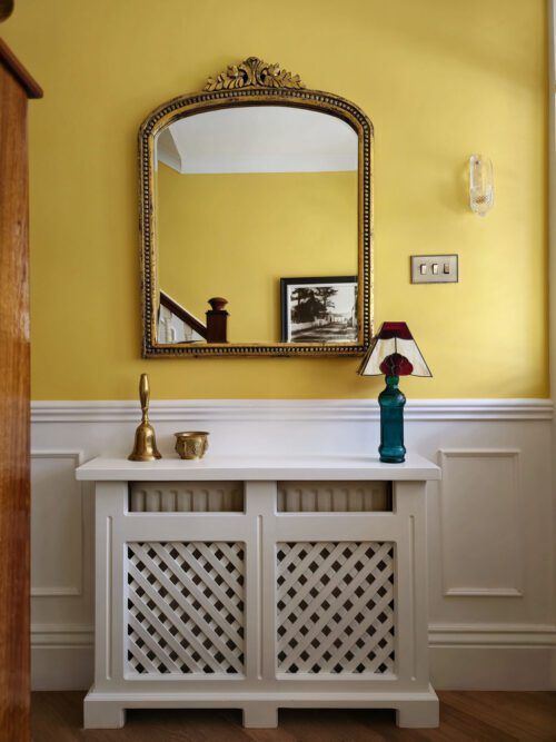 A hallway designed by Maureen Gomez Interiors, with a yellow wall and white panelling, featuring a decorative mirror with a gold frame above a white lattice radiator cover. On top sits a blue lamp with a red lampshade and a small brass candle holder. A framed black and white picture is in the background.