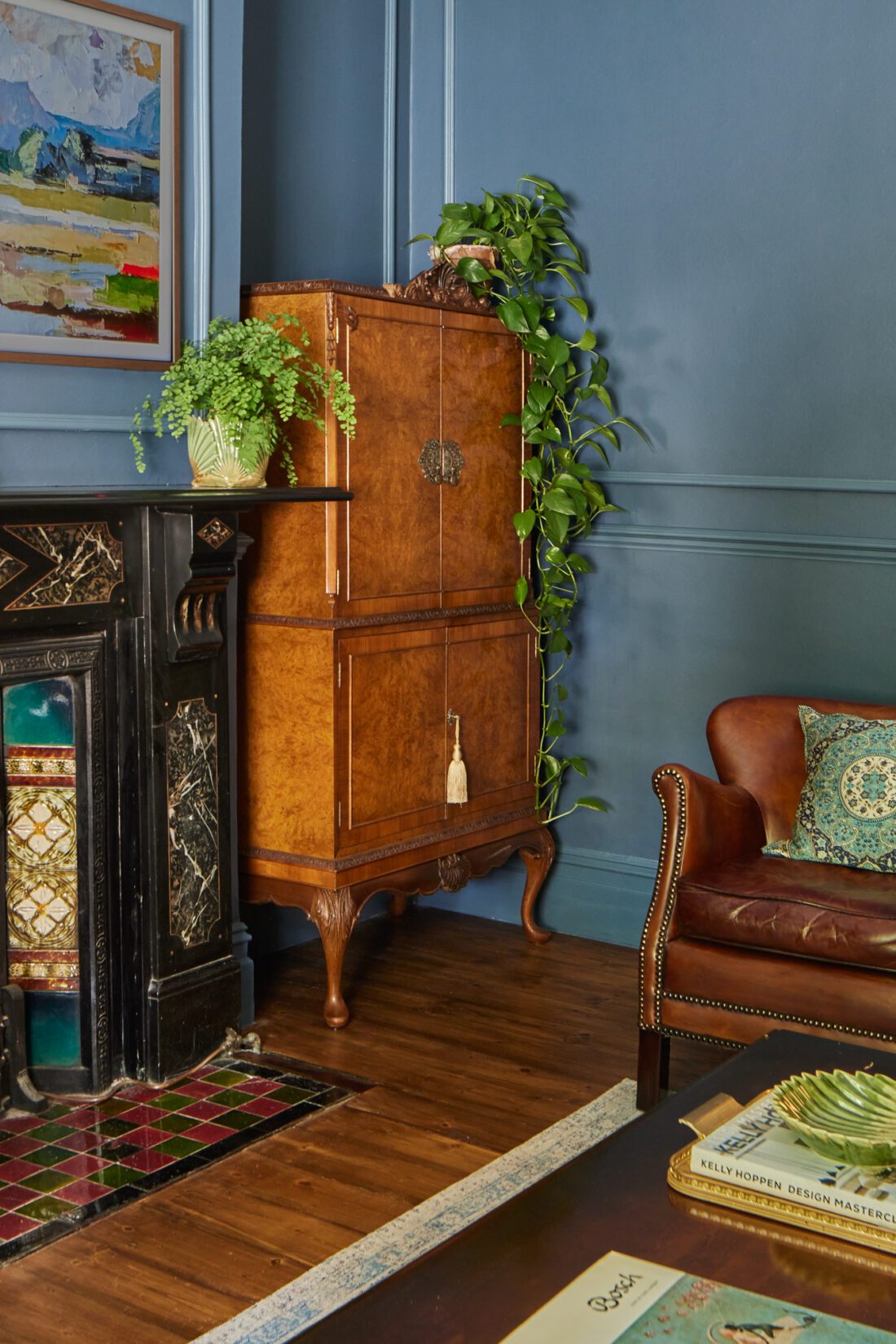 An elegant living room designed by Maureen Gomez Interiors, with an antique walnut cocktail cabinet adorned with trailing plants. An original Victorian fireplace with traditional tiles sits to the left. A leather armchair with a patterned cushion is beside a table holding books and a green plant.