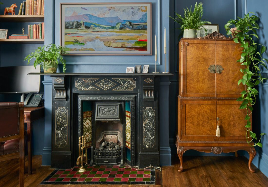 A cosy, elegant living room designed by Maureen Gomez Interiors, with a black marble fireplace surrounded by dark blue walls. A scenic painting hangs above, and nearby are lush green plants and an antique walnut cocktail cabinet. A hearth with red and green tiles lies on the wooden floor.