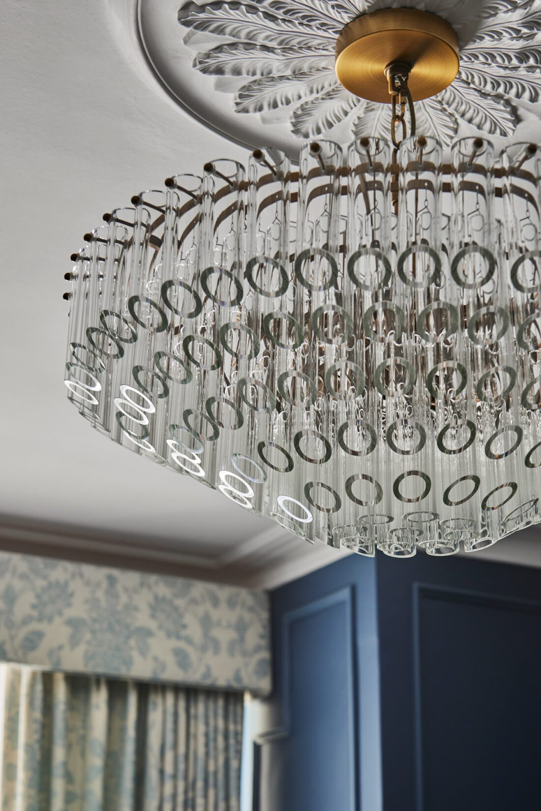 A luxurious mid-century style chandelier with circular crystal elements hangs from a ceiling that features an ornate, decorative medallion. The room has blue walls and patterned curtains, creating an elegant ambiance, designed by Maureen Gomez Interiors.