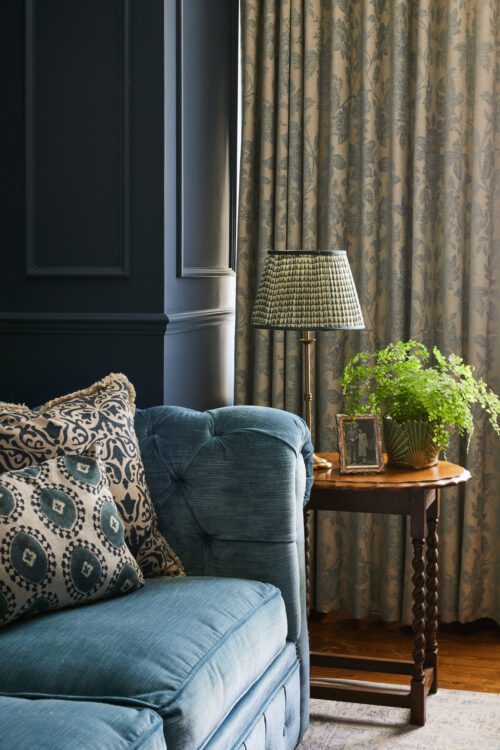 A cosy living room corner designed by Maureen Gomez Interiors, with a turquoise Chesterfield sofa adorned with patterned cushions. Beside it, a wooden table holds a brass lamp with a pleated patterned shade, a small fern plant, and a framed photo. Curtains with a subtle floral pattern hang in the background.
