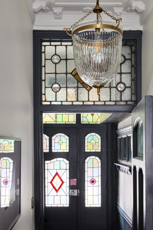 A Victorian hallway designed by Maureen Gomez Interiors, with stained glass windows and a door, featuring traditional colourful stained glass. A large decorative glass and brass pendant hangs from the ceiling. Walls are painted white with dark trim, and there's a small wall-mounted coat rack.