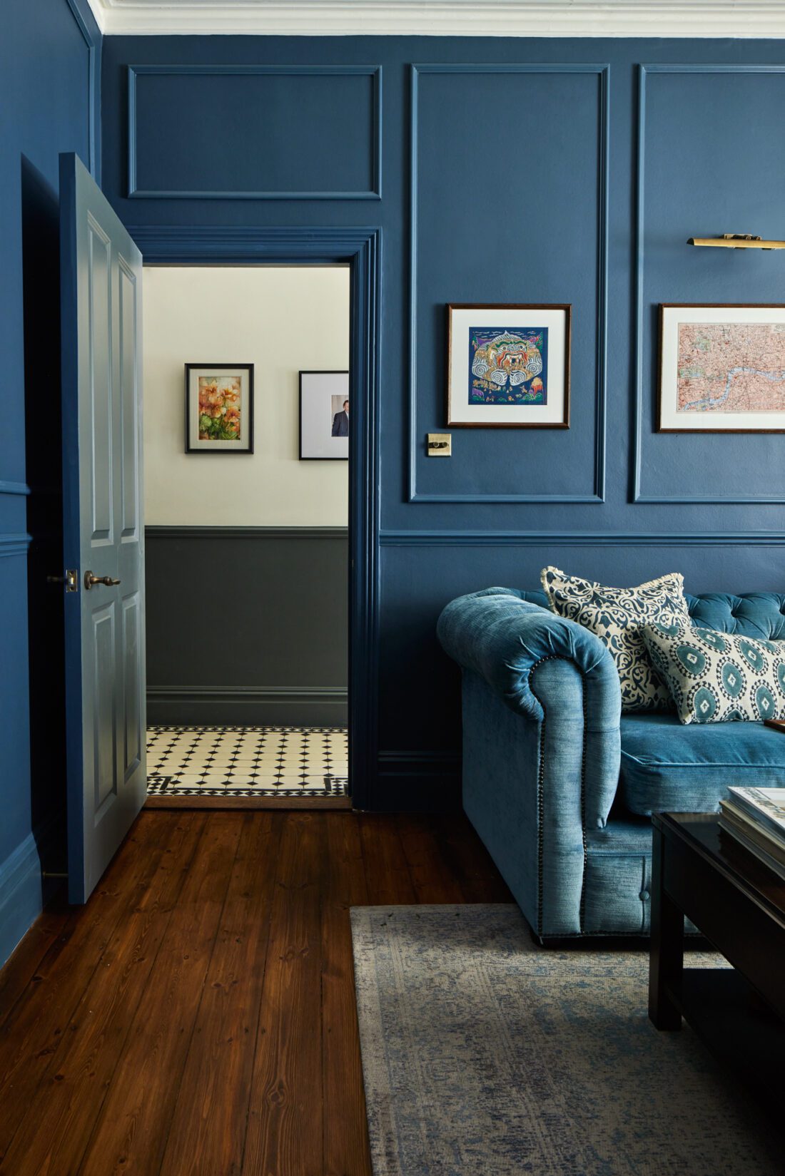 An elegant living room designed by Maureen Gomez Interiors, with dark blue panelled walls and a turquoise Chesterfield sofa adorned with patterned cushions. The room features framed artwork and wooden floors with a textured rug. An open door leads to a hallway with white and black tiled flooring.
