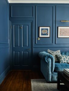 An elegant living room with dark panelled blue walls and door designed by Maureen Gomez Interiors. A plush turquoise chesterfield sofa with patterned cushions sits against the wall. Two framed artworks hang above the sofa. A dark wooden coffee table on a light rug completes the scene.