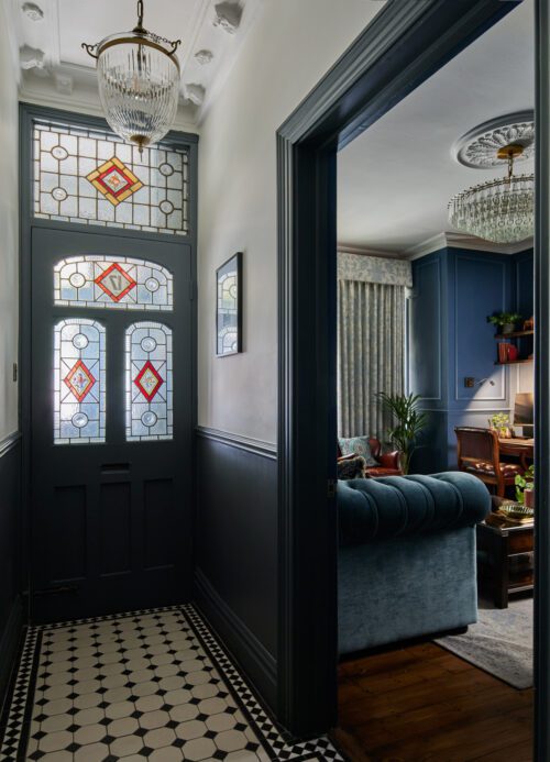 A hallway in Fulham designed by Maureen Gomez Interiors, boasts a stained glass front door with red diamond patterns. The floor features black and white tiles. To the right, an open door reveals a luxurious interior design with a cosy living room, blue sofa, ornate ceiling, and modern chandelier.