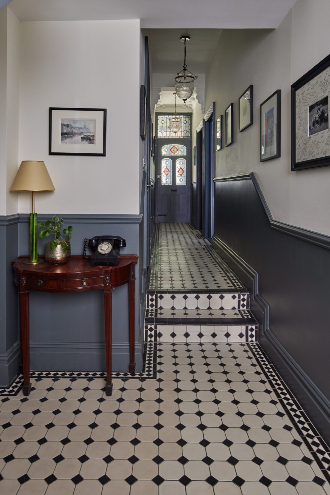 A Victorian hallway designed by Maureen Gomez Interiors, featuring a black and white tiled floor, a small antique wooden table with a lamp, potted plant, and rotary phone. Framed pictures adorn the walls, and stained glass decorates the door at the end.