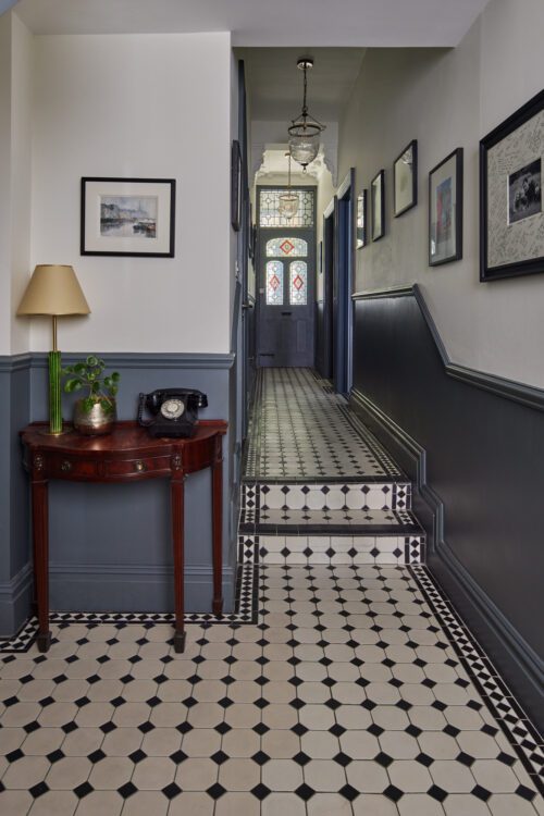 A Victorian hallway designed by Maureen Gomez Interiors, featuring a black and white tiled floor, a small antique wooden table with a lamp, potted plant, and rotary phone. Framed pictures adorn the walls, and stained glass decorates the door at the end.