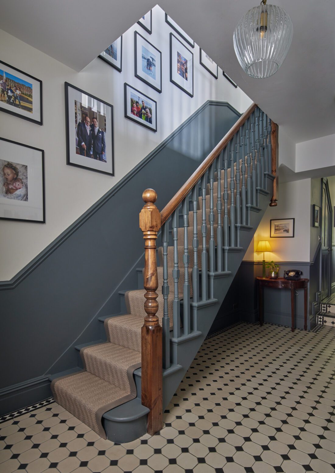 A traditional Victorian staircase with a wooden handrail and grey steps, designed by Maureen Gomez Interiors, featuring a beige carpet runner, is surrounded by framed photographs on the walls. The floor has a black and white hexagonal tile pattern. A vintage lamp sits on a small table nearby.