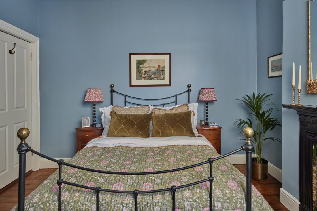 A cosy bedroom with blue walls designed by Maureen Gomez Interiors, features a black metal bed with a floral quilt. Two vintage wooden nightstands with patterned lamps and framed art hang above the bed. A potted plant and a dark fireplace complete the room’s decor.