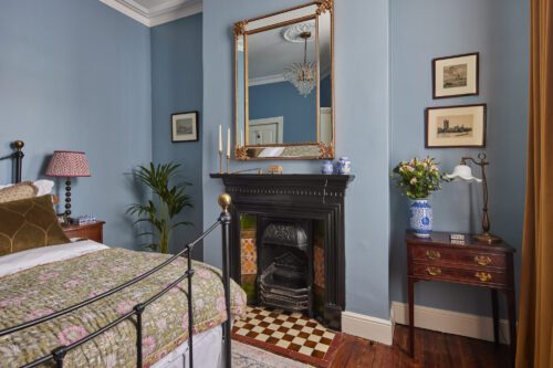 A cosy bedroom, designed by Maureen Gomez Interiors, with blue walls features a black iron bed, floral bedding, a Victorian fireplace with an antique mirror above, framed art, a small wooden side table with a lamp, and a potted plant. A chandelier hangs from the ceiling.