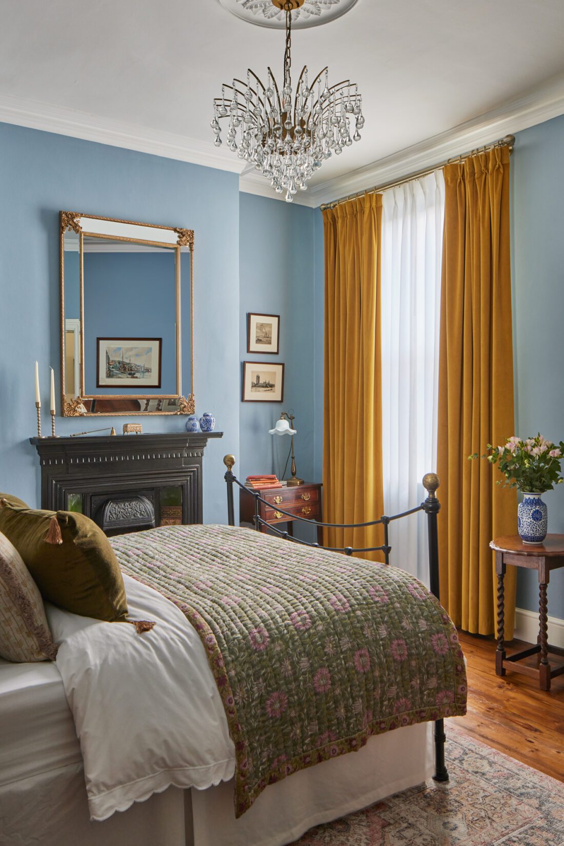 A cosy bedroom with blue walls designed by Maureen Gomez, features a black iron bed, a green floral quilt, and white pillows. An original Victorian fireplace with a mirror above, mustard curtains, a crystal chandelier, and a wooden side table with flowers complete the warm, elegant decor.